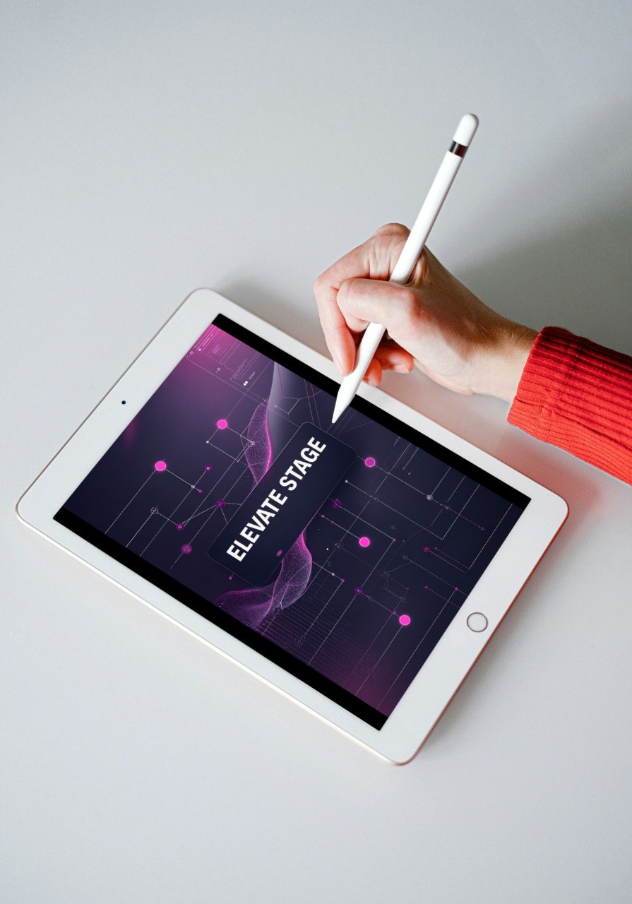 A white tablet on a light gray surface, with a hand holding a white stylus pointing at the screen. The screen displays a dark purple and pink digital interface with the text 'ELEVATE STAGE'.