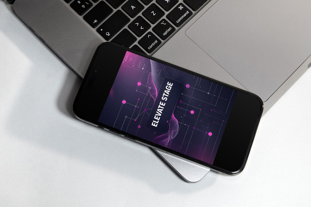 A black smartphone lying diagonally on top of a silver laptop, showing a purple and pink digital screen with the text 'ELEVATE STAGE'.